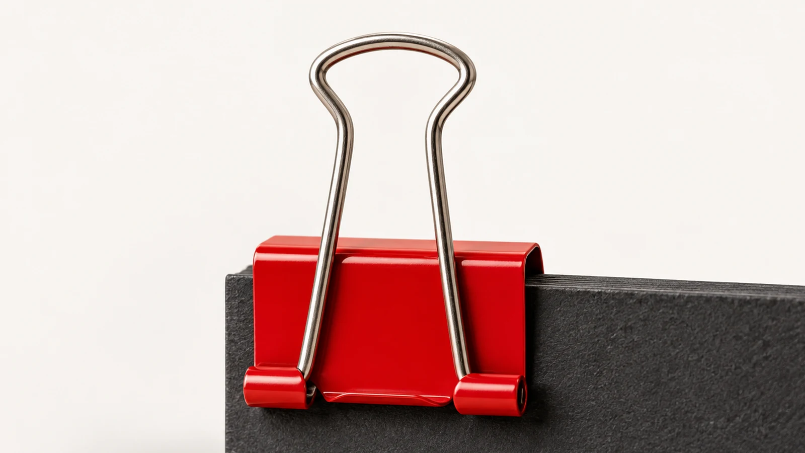 A red binder clip grips the edge of a charcoal folder against a smooth off-white background.