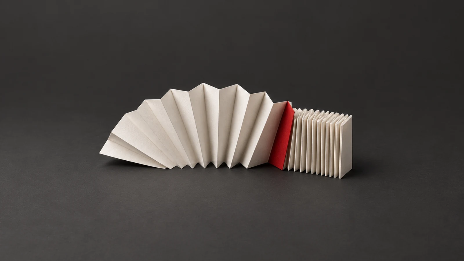 A partly opened off-white accordion strip meets a red fold and tightly packed unused pleats on charcoal.