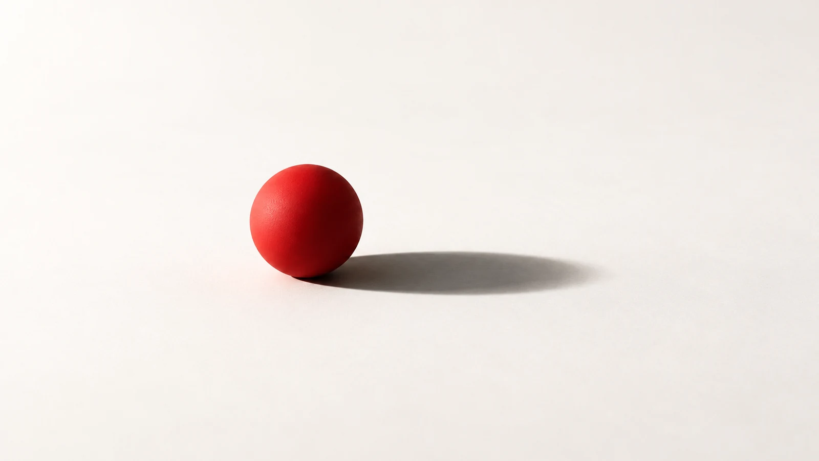 A small red sphere casts a much longer charcoal shadow across an off-white surface.