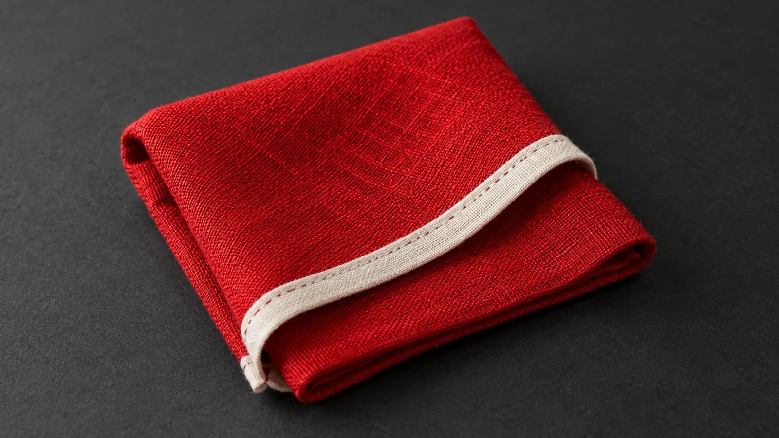 A folded red woven napkin shows an off-white stitched hem against a charcoal surface.